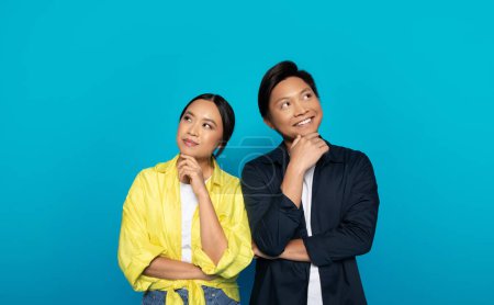 Photo for Positive pensive asian couple in casual look in different directions at empty space, isolated on blue studio background. Emotions think, sales choose, good news, question and dreams - Royalty Free Image
