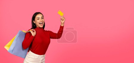 Foto de Mujer joven feliz emocionada sosteniendo bolsas de compras y demostrando tarjeta de crédito, Señora alegre recomendando pagos fáciles y servicios bancarios, posando aislado sobre fondo de estudio rosa, espacio de copia - Imagen libre de derechos