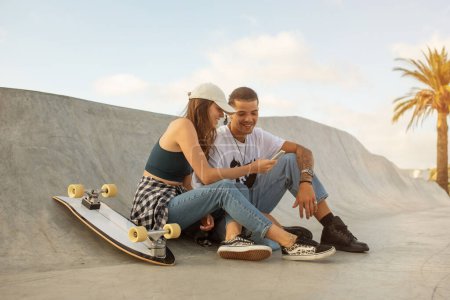Foto de Joven europeo hombre y mujer, skateboarders, tomar un descanso al aire libre, utilizando el teléfono celular. Sus monopatines sentados cerca, que representan un estilo de vida moderno y activo equilibrado con la interacción digital - Imagen libre de derechos