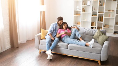 Photo for Indian couple finds comfort on cozy couch, leisurely using phone together, enjoying quiet, homey moments, browsing or communicating online, above view, free space - Royalty Free Image