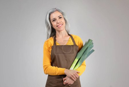 Photo for Smiling pretty calm senior woman farmer in apron hold onion leek, enjoy eco green harvest, isolated on gray background. Healthy food, agro business, farmer work, ad and offer - Royalty Free Image