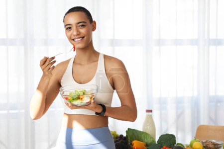 Photo for Smiling young african american woman in sportswear eat salad in kitchen light interior with fresh vegetables. Diet at home, weight loss, healthy food and health care, fit lifestyle - Royalty Free Image