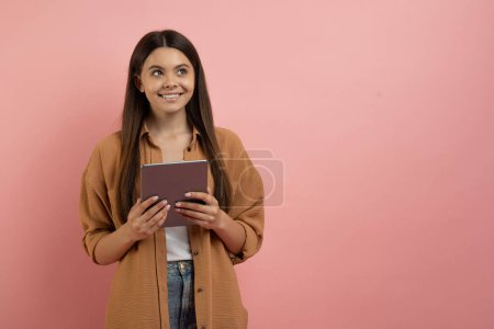 Téléchargez les photos : Jeune adolescente souriante tenant une tablette numérique et regardant loin, adolescente rêveuse pensive avec un gadget moderne dans les mains debout sur fond de studio rose, pensant à une offre agréable, Panorama - en image libre de droit