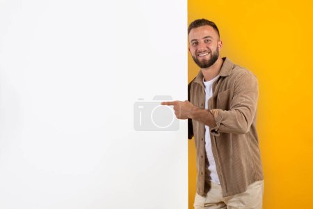Photo for Positive handsome young european man points fingers at big banner with free space, isolated on orange background, studio. Sale recommendation, advice for sale, ad and offer - Royalty Free Image