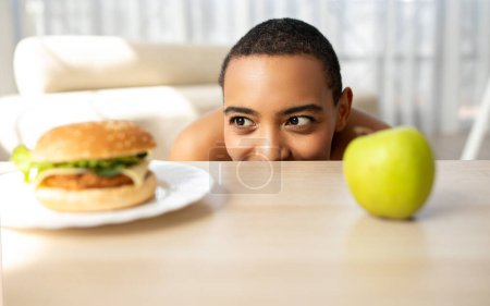 Foto de Alegre divertido joven afroamericano dama mirar hamburguesa en la mesa con manzana verde en el interior de la cocina, de cerca. Elija comida sana o rápida en el hogar, atención médica, pérdida de peso - Imagen libre de derechos