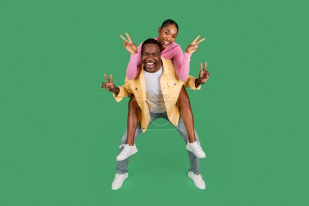 Photo for Cute happy beautiful millennial african american couple have fun on green studio background. Positive pretty young black woman piggybacking her boyfriend, showing peace gesture and smiling - Royalty Free Image