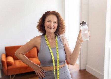 Téléchargez les photos : Concept de perte de poids. Femme âgée sportive avec ruban à mesurer tenant une bouteille d'eau et souriant à la caméra, heureuse femme âgée recommandant une boisson saine tout en suivant un régime, debout à l'intérieur de la maison - en image libre de droit