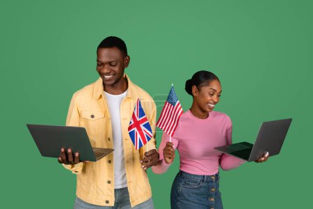 Téléchargez les photos : Heureux homme et femme noirs millénaires tenant des ordinateurs portables et des drapeaux du Royaume-Uni et des États-Unis, fond de studio vert. Étudiants afro-américains apprenant l'anglais langue étrangère en ligne. Immigration, e-éducation - en image libre de droit
