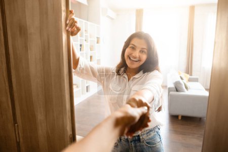 Foto de Mujer india graciosa se encuentra junto a la puerta principal abierta, extendiendo el cálido apretón de manos a los huéspedes, invitando a su acogedora casa con expresión amistosa y acogedora - Imagen libre de derechos