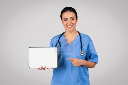 Photo for Professional latin woman doctor in blue uniform confidently points towards a digital tablet with blank screen, offering space for customizable content against grey studio background - Royalty Free Image