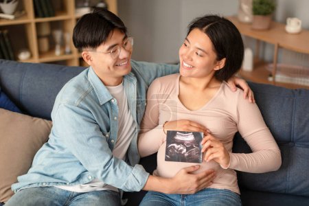 Téléchargez les photos : Grossesse. Couple coréen souriant attend bébé, posant tenant photo échographie près du ventre des femmes enceintes, étreignant assis sur le canapé à la maison. Nouvelle vie, concept de famille et d'accouchement - en image libre de droit