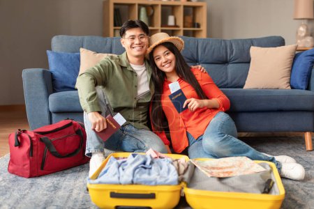 Téléchargez les photos : Des vacances. Couple de voyageurs asiatiques excités à la maison, tenant des billets de voyage et des passeports près d'une valise ouverte, assis sur le sol se préparant pour les voyages de vacances, souriant à la caméra - en image libre de droit