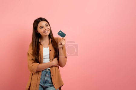 Foto de Chica adolescente soñadora sosteniendo tarjeta de crédito y mirando el espacio de copia, feliz adolescente sonriente disfrutando de libertad financiera y servicios bancarios de calidad, de pie sobre fondo de estudio rosa - Imagen libre de derechos