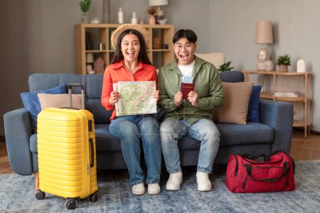 Foto de Alegre pareja de viajeros japoneses celebrando viaje de vacaciones sentado con mapa de viajes y entradas en casa, posando con equipaje, mostrando sus pasaportes con tarjeta de embarque en emoción - Imagen libre de derechos