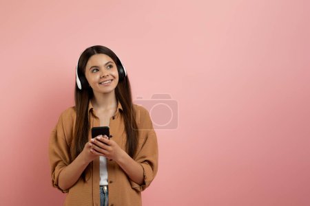 Foto de Sonriente chica adolescente con auriculares sosteniendo el teléfono inteligente y mirando el espacio de copia sobre el fondo del estudio rosa, sonriente adolescente femenina disfrutando de los servicios de transmisión para escuchar música, Panorama - Imagen libre de derechos
