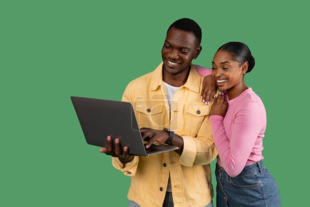 Téléchargez les photos : Aimant jeune homme et femme afro-américain joyeux embrassant et utilisant un ordinateur portable isolé sur fond de studio vert, espace de copie. Noir couple shopping, bancaire en ligne, à la recherche de belle film - en image libre de droit