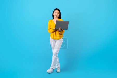Photo for Positive cheerful beautiful young asian lady in casual outfit posing on blue studio background, holding laptop computer. Happy chinese woman freelancer working or shopping online, copy space - Royalty Free Image