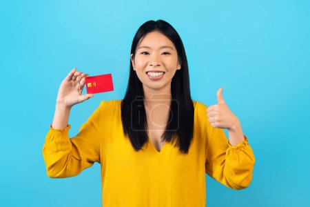 Foto de Alegre bastante millennial asiático mujer con frenillos y en casual mostrando rojo tarjeta de crédito y pulgar hacia arriba en la cámara, sonriendo, recomendando el pago sin contacto, fácil de banca rápida, azul fondo de estudio - Imagen libre de derechos