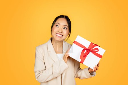 Photo for Cheerful Asian millennial woman amidst celebration, holding carefully wrapped gift with excitement. The ambiance is festive and her demeanor is imbued with warm, generous spirit of holiday giving - Royalty Free Image