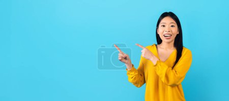 Téléchargez les photos : Enthousiaste jolie femme asiatique millénaire portant chemise jaune pointant vers l'espace de copie pour la publicité ou le texte isolé sur fond bleu studio, panorama. Excellente offre, bonne affaire - en image libre de droit