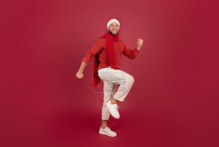 Photo for Smiling millennial caucasian man in Santa hat walk, enjoy holidays, spare time, has fun, isolated on burgundy studio background, full length. Christmas, New Year emotions, lifestyle - Royalty Free Image