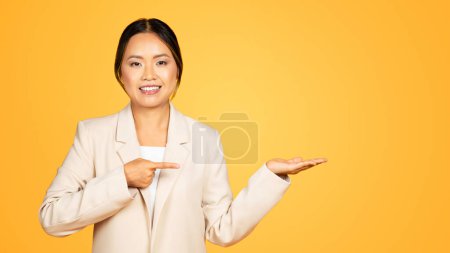 Téléchargez les photos : Femme asiatique souriante, doigt millénaire pointé, attirant l'attention sur la quantité d'espace libre, isolée sur fond jaune, studio, panorama. Permet l'utilité polyvalente dans la création de contenu - en image libre de droit