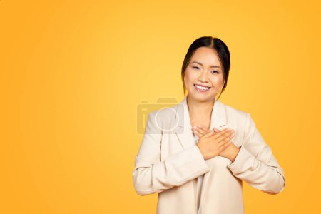 Foto de Momento sentido de sonriente mujer milenaria asiática coloca las manos en el pecho, los ojos relucientes con genuina gratitud y amabilidad, aislados en el fondo del estudio naranja. Personas emociones - Imagen libre de derechos