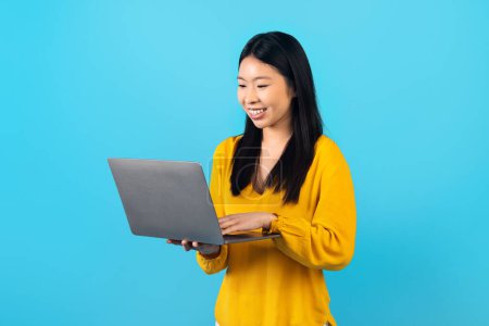 Photo for Cheery attractive young korean woman wearing yellow shirt using laptop computer isolated on blue studio background. Asian lady digital nomad shopping or studying online, have remote job - Royalty Free Image