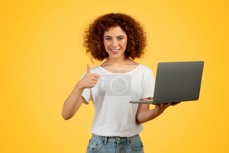 Téléchargez les photos : Femme adolescente confiante et souriante travaillant sur un ordinateur portable, doigt levé dans un moment de pensée expresse, fusionnant des mondes de vitalité et de travail concentré, isolée sur fond jaune. Recommandation, approbation - en image libre de droit