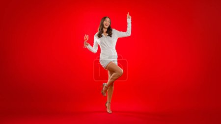 Photo for Holiday party. Full length of happy woman in cocktail dress holding champagne glass and dancing while posing over red studio background. Birthday, bachelorette and new year celebration. Panorama - Royalty Free Image