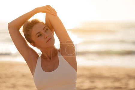 Photo for Active young European woman deeply stretching her arms, practicing yoga, with the gentle sea waves caressing the sandy beach in the sunset time - Royalty Free Image