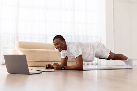 Photo for Happy young black man in sportswear doing workout, using laptop, having online fitness class from home, living room interior, copy space. Sporty african american guy planking, watching video - Royalty Free Image