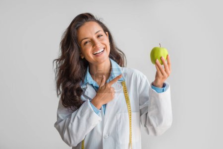 Téléchargez les photos : Joyeux nutritionniste tenant une pomme verte fraîche à la main et la pointant du doigt, debout avec un ruban adhésif drapé autour du cou, soulignant l'importance d'une alimentation saine et de mesures - en image libre de droit