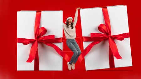 Photo for Christmas Gifts Offer. Joyful young lady in Santa hat posing with two huge Xmas presents boxes with ribbon bows over red studio background. Winter season holidays and sales. Panorama - Royalty Free Image