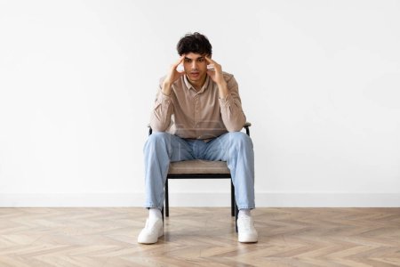 Photo for Concerned Middle Eastern Guy Thinking And Worrying About Problems Touching Head Sitting In Chair Over White Wall Background Indoors. Depression And Male Mental Health Issue, Stressful Life - Royalty Free Image