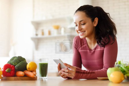 Photo for Weight Loss. Smiling Lady In Sportswear Using Phone Application Leaning On Dining Table With Vegetables In Modern Kitchen Indoor, Browsing Healthy Low Calories Recipes Online - Royalty Free Image