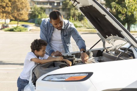 Photo for Black father and preteen son checking car engine outdoors. African american dad and school aged kid standing next to open automobile hood, fixing broken auto, traveling together - Royalty Free Image