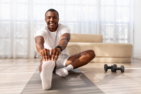 Photo for Morning routine. Athletic millennial black man warming up and stretching, preparing for workout with dumbbells, training on mat in living room interior at home, free space. - Royalty Free Image