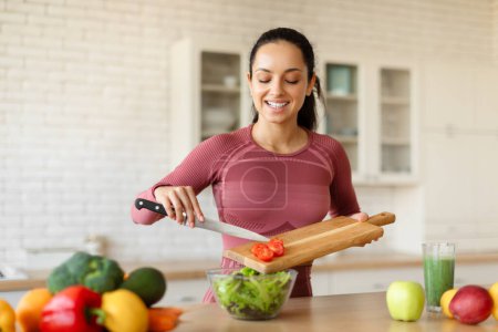 Photo for Happy fit woman cooking healthy vegetable salad, cutting tomatoes on wooden board, preparing food for slimming in kitchen interior. Young woman making dinner for weight loss dieting - Royalty Free Image