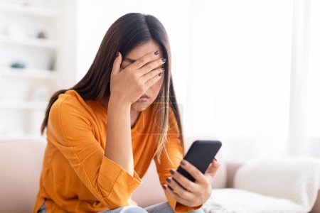 Photo for Bad News Concept. Upset Confused Woman Holding Smartphone, Covering Face with Palm, Sad Adult Asian Lady Reading Unpleasant Message Sitting On Couch At Home, Copy Space - Royalty Free Image