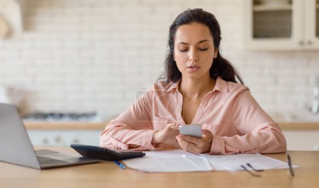 Photo for Concentrated woman sitting at wooden table, holding smartphone with laptop and paperwork in front of her, set against modern kitchen backdrop, panorama, free space - Royalty Free Image