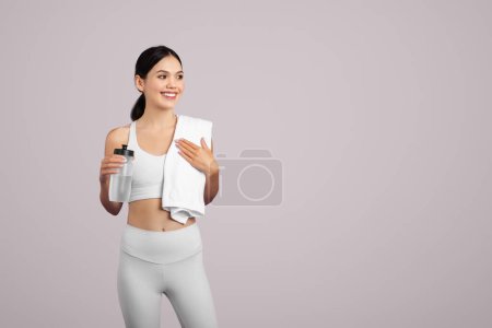 Foto de Ajuste mujer europea en ropa deportiva sosteniendo una botella de agua y drapeando una toalla sobre su hombro, señalando post-entrenamiento sobre un fondo claro, espacio libre - Imagen libre de derechos