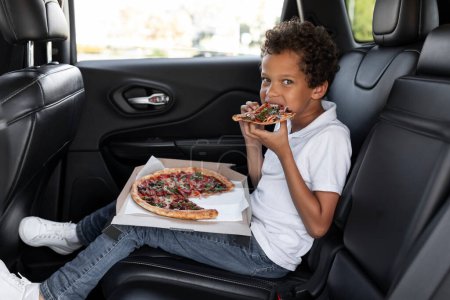 Photo for Hungry african american boy eating pizza while riding car. Happy cute curly black school aged kid sitting at auto backseat, enjoying his lunch, looking at camera, copy space - Royalty Free Image
