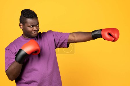 Photo for Black boxer guy throwing punch wearing red boxing gloves, looking aside standing over yellow background. Sportsman having box training in studio. Sport and workout concept - Royalty Free Image