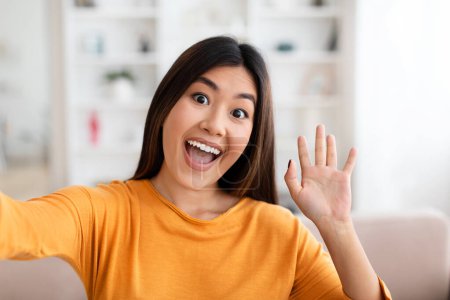 Téléchargez les photos : Belle jeune femme asiatique positive à manches longues orange prenant selfie à la maison, agitant joyeusement et souriant à la caméra, influenceur montrant son nouvel appartement. Concept de blogging - en image libre de droit