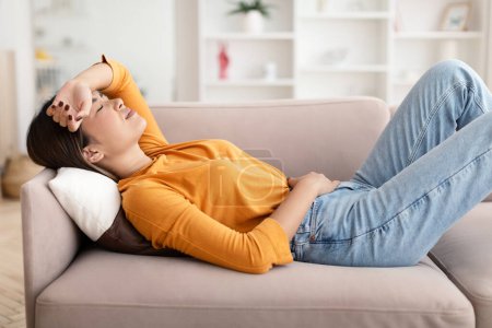 Photo for Feeling unwell, menstruation, flu, migraines and illness. Sad despair young asian woman lying on couch suffering from headache and abdominal pain in living room home interior - Royalty Free Image