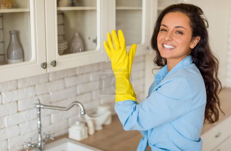 Téléchargez les photos : Femme rayonnante en chemise bleu clair, posant en toute confiance dans la cuisine moderne, portant des gants jaunes, prête pour le nettoyage, debout dans l'intérieur de la cuisine et souriant, espace libre - en image libre de droit