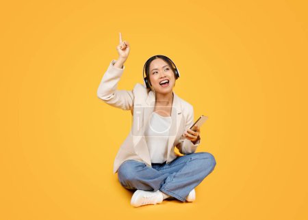Photo for Cheerful young korean lady in wireless headphones with phone has fun, singing song, sit on floor, isolated on yellow studio background. Emotions, relax with music, spare time - Royalty Free Image