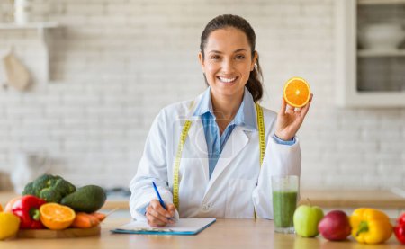 Foto de Señora caucásica en una bata de laboratorio presentando una naranja mientras escribe notas sentadas en la cocina moderna, simbolizando la orientación nutricional, mirando y sonriendo a la cámara - Imagen libre de derechos