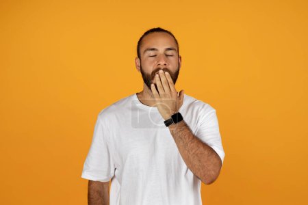 Téléchargez les photos : Fatigué jeune homme européen en t-shirt blanc couvre la bouche avec sa main et bâillements, isolé sur fond orange. Surmenage, ennui, mode de vie, temps de dormir et de se réveiller tôt - en image libre de droit
